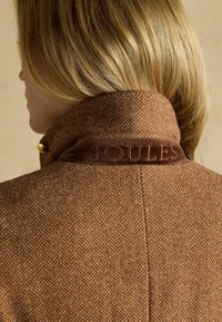 Sacou cu model în herringbone de culoare brună, cu un guler din corduroy brun și butoane aurii. Dispune de logo-ul gravat "JOULES" pe guler.