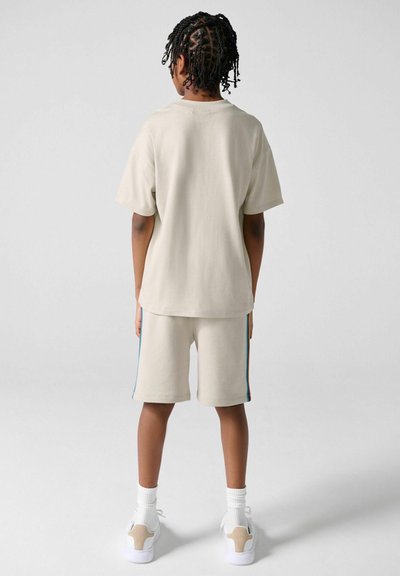 Barn med fletninger iført beige oversized T-shirt og matchende shorts med sidestriber, hvide sokker og hvide sneakers, stående med ryggen mod kameraet.