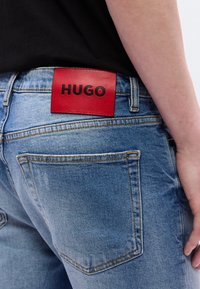 Gros plan d'une personne portant un jean bleu avec une étiquette en cuir rouge affichant le mot HUGO cousu au-dessus de la poche arrière.