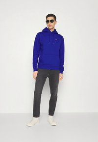 Felpa con cappuccio blu reale in cotone, caratterizzata da una tasca frontale, polsini a costine e un piccolo logo sul petto, abbinata a jeans scuri e scarpe bianche.