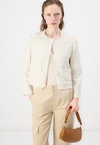 Kreemi tweed takki, jossa on hapsuilla viimeistelyt, kaksi etutaskua; päällä beigepinkki toppi ja yhdistettynä vaaleanruskeisiin housuihin sekä pieneen ruskeaan käsilaukkuun.