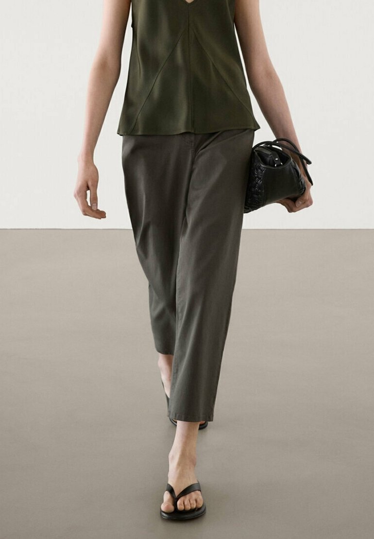 Personne portant un haut sans manches vert olive, un pantalon sombre ample, des sandales noires et un sac à main noir, marche sur un sol uni.