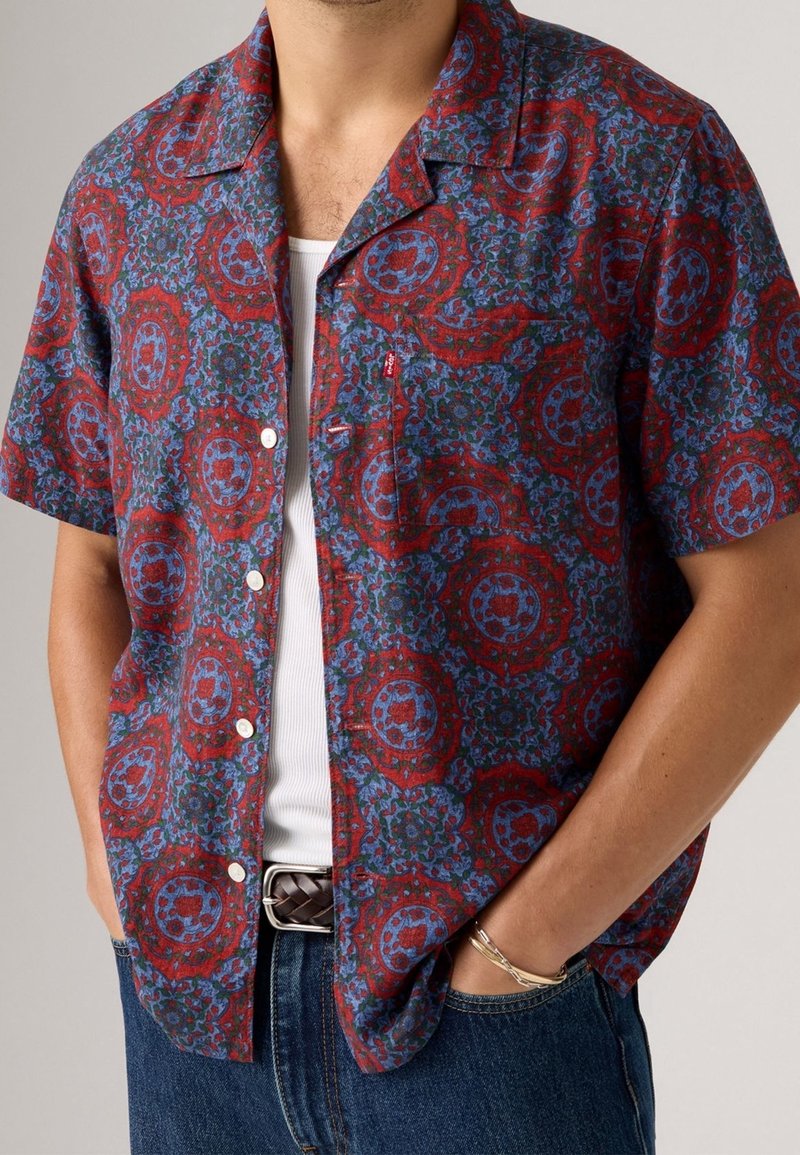 Koszula z krótkim rękawem w czerwono-niebieski kwiatowy wzór, wykonana z lekkiego materiału, z kołnierzem z wycięciem i białymi guzikami.