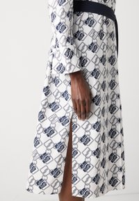 Vestito lungo bianco con un motivo a catena navy, dotato di uno spacco laterale e maniche lunghe. Presenta una cintura navy a contrasto in vita.