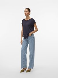 Žena oblečená v námořnickém krátkém rukávu, světle modrých rovně střižených džínech a olivových lodičkách s špičatou špičkou, stojí s jednou rukou v kapse.