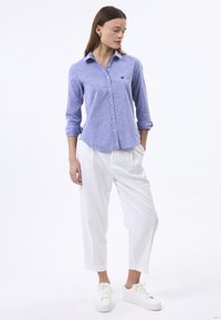 Camicia azzurra chiara con una texture sottile, maniche lunghe arrotolate, abbinata a pantaloni bianchi a taglio largo e sneakers bianche.