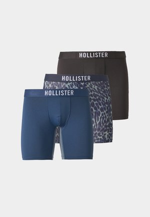 Junger Mann steht barfuß in schwarzen Hollister Boxershorts vor einem schlichten hellen Hintergrund.