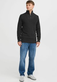 Dunkelgrauer Strickpullover mit halbem Reißverschluss und strukturiertem Gewebe, mit gerippten Bündchen und Saum, getragen mit blauen Jeans und grauen Sneakern.