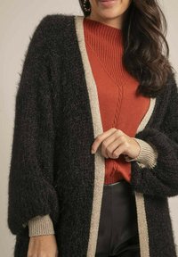 Cardigan noir en peluche avec des bordures crème, porté sur un pull côtelé couleur rouille. Il présente des manches bouffantes et de subtils détails en tricot torsadé.