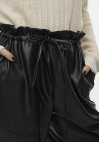 Zwarte leren broek met een hoge tailleband, voorzien van een gerimpelde rand en een gestrikte koord. Aan beide zijden zijn zakken zichtbaar.