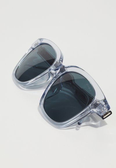 Transparent gray sunglasses with dark lenses, angular design, bold frame, and subtle hardware accents.