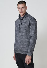 Sudadera gris con una superficie texturizada, cuello alto y cierre de cordón. Presenta puños y bajo acanalados, con un patrón sutil en toda la prenda.