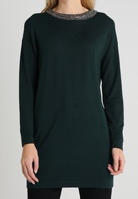 Tunica verde a maniche lunghe realizzata in tessuto morbido, con scollatura rotonda decorata da dettagli elaborati, caratterizzata da una vestibilità ampia e un taglio dritto.