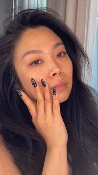 Långa, spetsiga naglar med svart nagellack prydda med vita prickar; vågigt mörkt hår; neutral hudton; mjukt ljus.