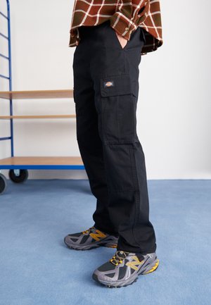 Person, die schwarze Cargohosen, graue und gelbe Turnschuhe sowie ein braunes kariertes Hemd trägt und neben einer blauen und hölzernen Regaleinheit steht.