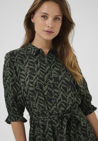 Robe chemise à motif vert avec fermeture à boutons, manches courtes bouffantes et ceinture à nouer. Le tissu présente un design de feuilles sombres sur un fond noir.