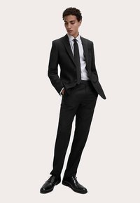 Černý na míru šitý oblek s dvouknoflíkovým sakem a kalhotami s rovnými nohavicemi. Kombinováno s bílou košilí a černou kravatou. Dokončeno lesklými černými botami.