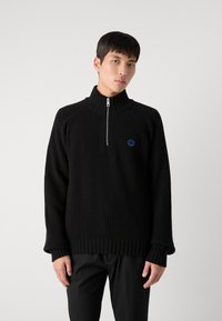 Maglione nero a costine con alto colletto, chiusura a metà zip e logo circolare blu sul petto. Presenta maniche lunghe e larghe e polsini a costine.