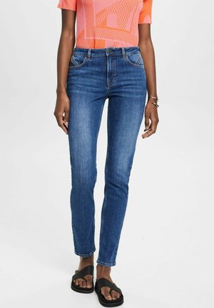 Femme portant un jean skinny bleu taille mi-haute, un top corail à manches courtes, des sandales noires et des bracelets argentés sur fond blanc.
