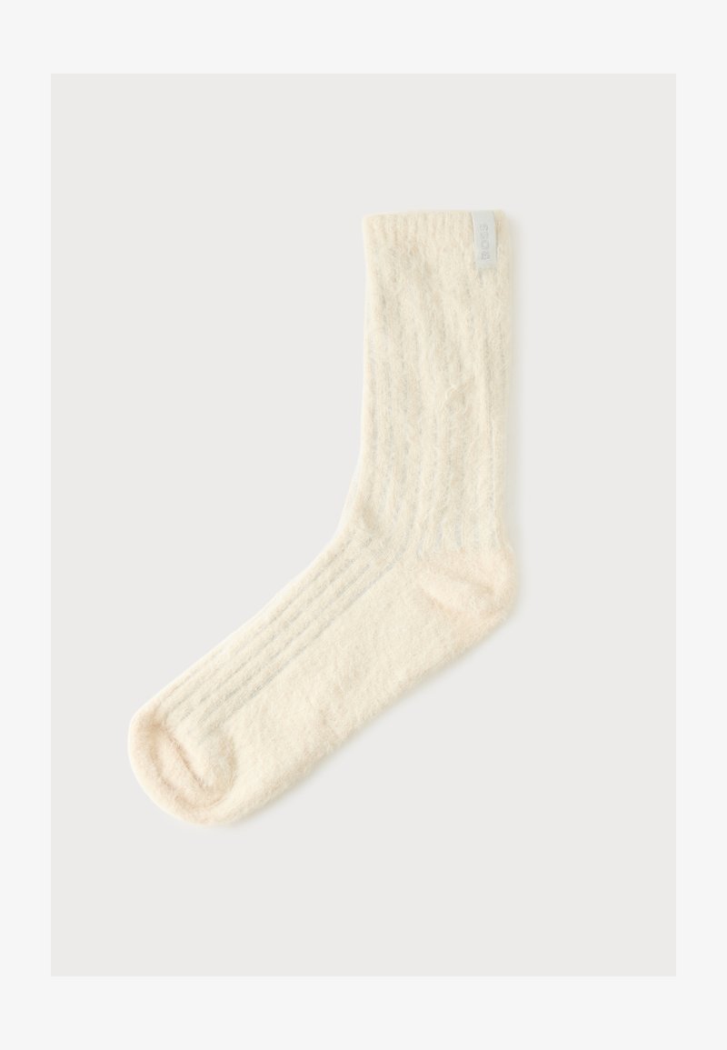 Chaussette tricotée texturée couleur crème unique avec une petite étiquette en tissu près du bord supérieur sur un fond clair.