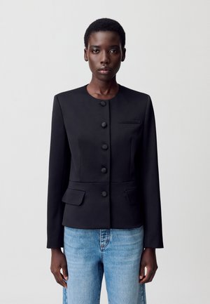 Modèle portant une veste noire sur mesure avec des boutons et des poches, associée à un jean bleu clair, sur fond blanc uni.