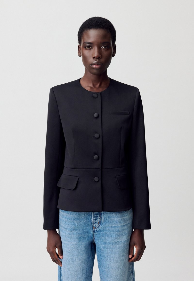 Modelo vistiendo un blazer negro a medida con botones y bolsillos, combinado con jeans azul claro, sobre un fondo blanco liso.