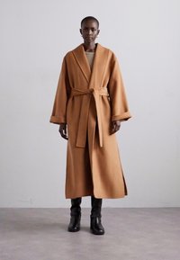 Palton lung, de culoare cămilă, realizat dintr-un material moale și texturat, cu un design de tip wrap, mâneci ample și o curea asortată la talie.