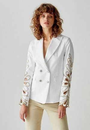 Blazer bianco doppiopetto con ritagli decorativi sulle maniche e dettagli in marrone chiaro. Tessuto liscio con design aderente e leggera lucentezza.