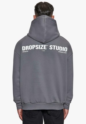 Homme aux cheveux courts et foncés portant un sweat à capuche gris avec "DROPSIZE STUDIO" et un petit texte blanc imprimé dans le dos.