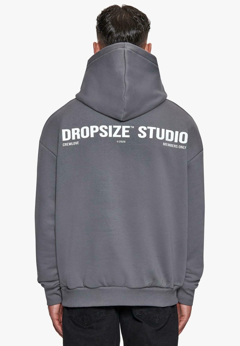 Homme aux cheveux courts et foncés portant un sweat à capuche gris avec "DROPSIZE STUDIO" et un petit texte blanc imprimé dans le dos.