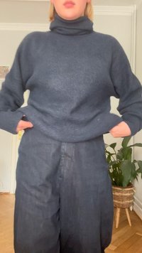 Suéter de gola alta cinza escuro em malha suave e texturizada, combinado com calças largas escuras à medida. Bolso visível em ambas as peças.