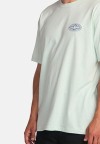 Lys mintgrøn bomulds T-shirt med korte ærmer, der har et lille blåt logo på brystet. Glat tekstur og afslappet pasform.