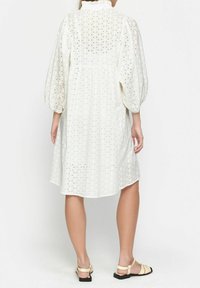 Vestido blanco con un patrón texturizado de ojales y mangas abullonadas, que presenta un cuello alto y un ajuste relajado que cae a mitad del muslo.
