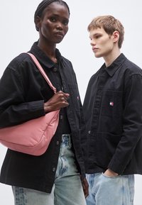 Schwarze Baumwolljacken an zwei Modellen, eines hält eine rosa Umhängetasche. Jeans mit verwaschener Textur. Die Jacken sind mit Knopfverschlüssen und Taschen versehen.