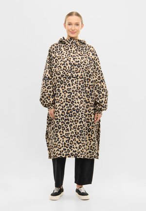 Kvinde iført en løs leopardmønstret hættetrøje over sorte bukser med vide ben og sort-hvide sneakers, stående mod en ensfarvet baggrund.