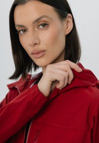 Czerwona wodoodporna kurtka z kapturem, z zapięciem na zamek i elastycznymi mankietami. Gładki, teksturowany materiał widoczny w zbliżeniu.