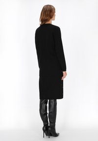 Rochie neagră cu mâneci lungi, cu o croială dreaptă, având accente cu nasturi laterali și o textură moale, asortată cu cizme negre lucioase până la coapse.