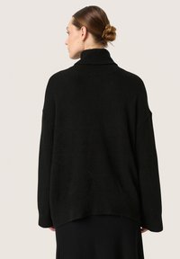 Melna pārstrādāta turtlenecks džemperis ar strukturētu apdari, kritieniem pleciem un garāku aizmugurējo malu. Krāsa ir vienkārša melna bez rakstiem.