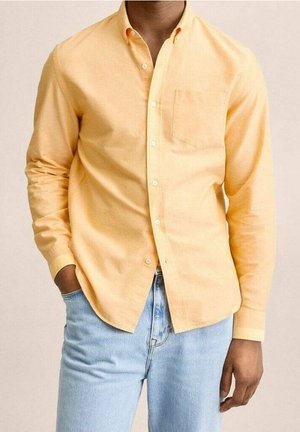 Mann trägt ein hellorangenes, langärmeliges Hemd mit Brusttasche und hellblaue Jeans, Hände entspannt seitlich.