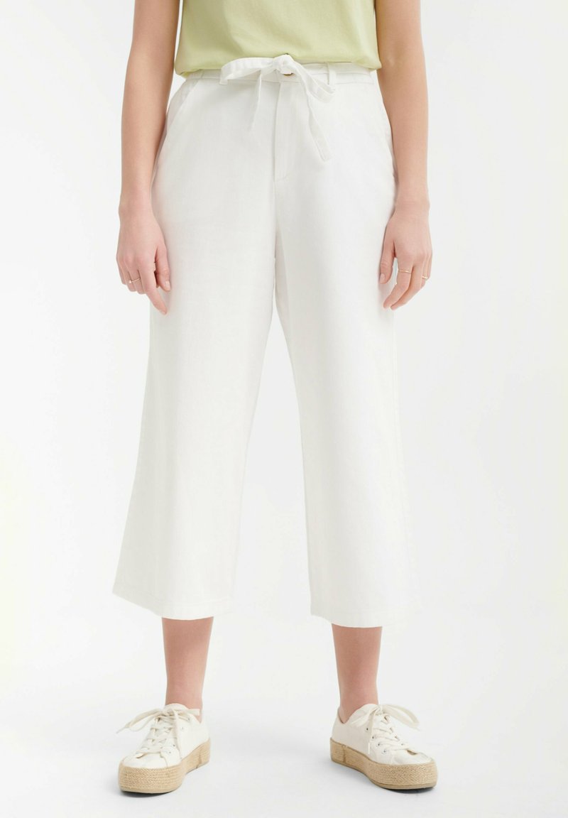 Pantalones culotte de lino blancos con cintura anudada, que presentan un diseño de pierna ancha. Combinados con unas zapatillas blancas y una camisa verde claro.