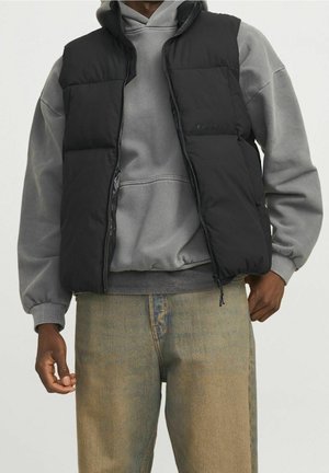 Person iført sort dynejakke over en grå hættetrøje og lyseblå jeans, vist fra skuldre til knæ mod en ensfarvet baggrund.