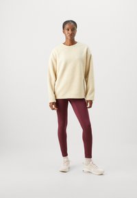 Felpa in pile color crema con vestibilità rilassata, abbinata a leggins bordeaux e sneakers chiare. Texture liscia e design semplice.