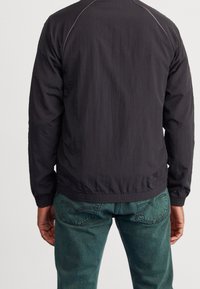 Black lightweight jacket with ribbed cuffs and hem, featuring a smooth texture and contrast stitching. Paired with green denim jeans.
