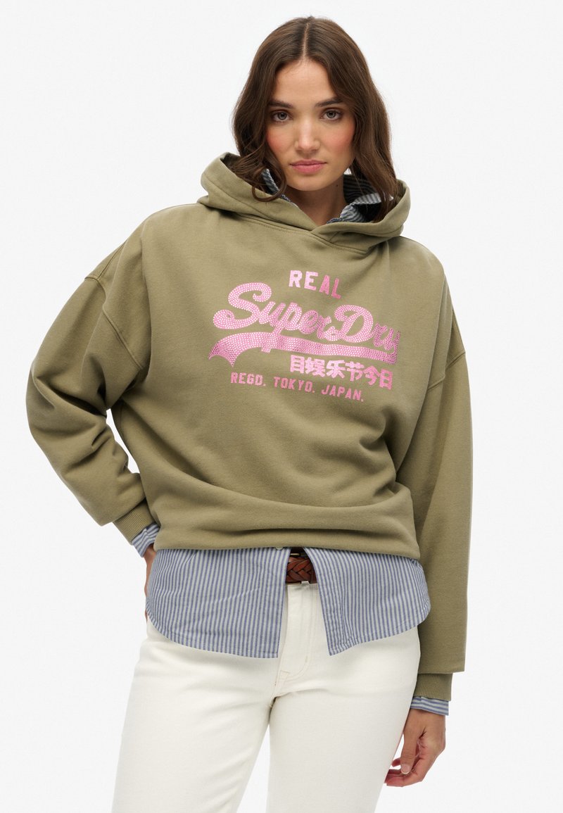 Felpa oversize verde oliva con testo grafico rosa, indossata sopra una camicia a righe blu e bianca. Pantaloni bianchi e una cintura marrone sono visibili.