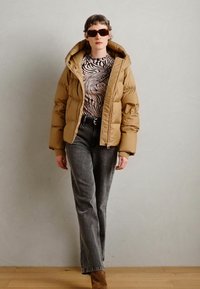Veste puffer kaki avec capuche, haut à motifs noir et blanc, jeans gris et bottines en suède marron. Des lunettes de soleil carrées complètent la tenue.
