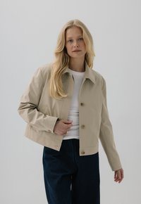 Beżowa krótka kurtka z kołnierzem, dwiema guzikami i bocznymi kieszeniami, zestawiona z ciemnoniebieskimi spodniami w szerokim kroju. Widoczny gładki materiał.