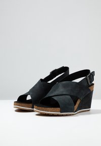 Sandales compensées noires avec semelles en liège, brides croisées et boucles arrière ajustables sur une surface blanche avec un fond gris clair uni.