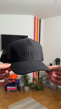 Svart snapback-keps med texturerat tyg, med en upphöjd "HUGO"-logotyp i svart och färgstänk genom hela materialet.