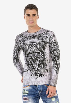Langärmliges Hemd in Grau mit einem schwarzen Grafikdesign, das Schlangen und kunstvolle Muster zeigt, kombiniert mit abgenutzten blauen Jeans.