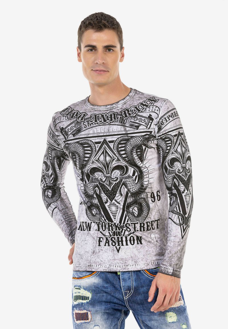 Langärmliges Hemd in Grau mit einem schwarzen Grafikdesign, das Schlangen und kunstvolle Muster zeigt, kombiniert mit abgenutzten blauen Jeans.